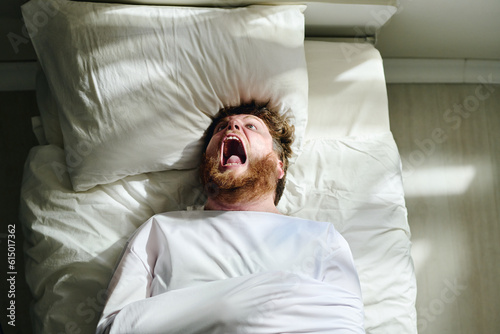Young insane man with psychosis in straitjacket shouting while lying on bed in mental hospital during therapy recommended by his doctor