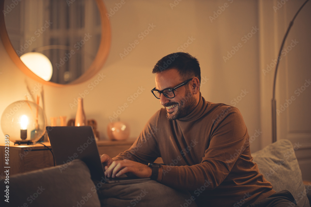 Fototapeta premium Man using a laptop, sitting on the sofa, enjoying himself at home, night time.