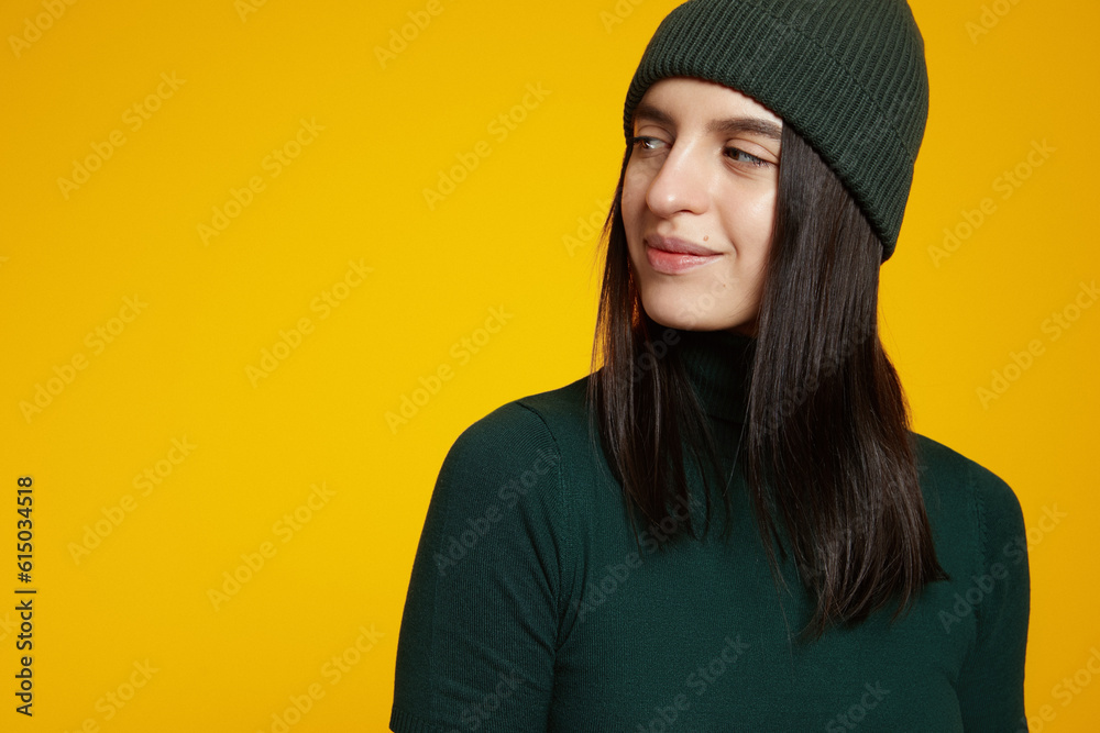 Close up portrait of young attractive woman in green hat and t-shirt, looking at free empty space for text, isolated over yellow studio background