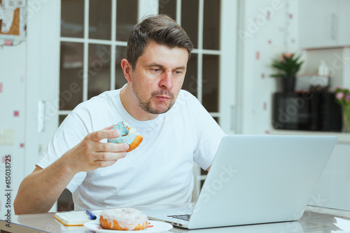 Freelancer man eating unhealthy junk food while working from home. Attractive handsome young male feeling happy and enjoy, use laptop computer study learn online and eat donut in the kitchen