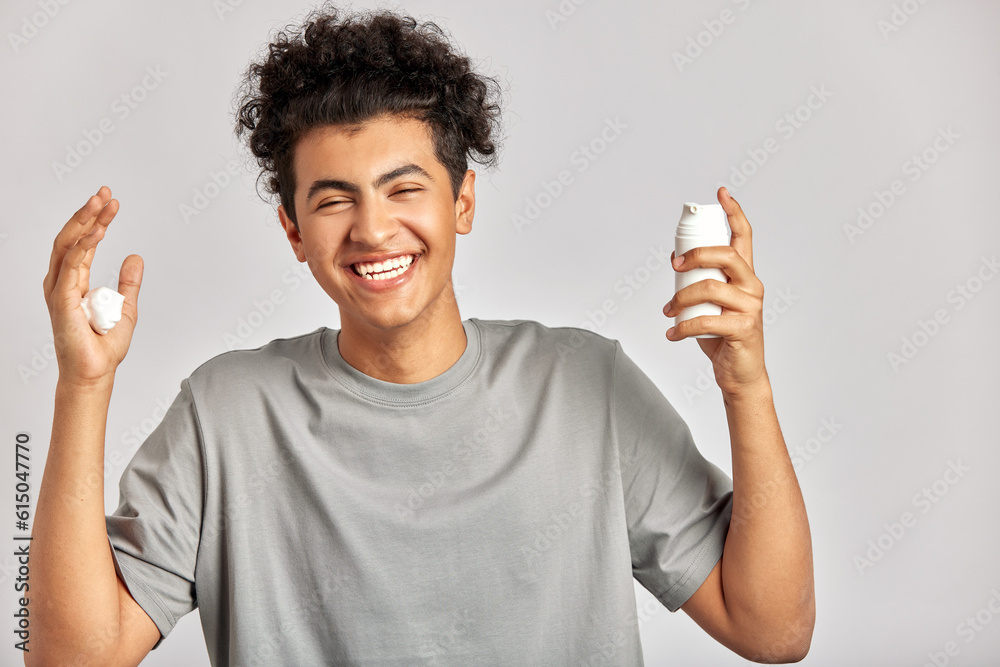 Young handsome smiling guy with black curly hair squeezing skincare cream from the dispenser to his hand. Biracial man in studio with healthy skin procedure. Wellness, cosmetics and skincare for men. Young handsome smiling guy with black curly hair squeezing skincare cream from the dispenser to his hand. Biracial man in studio with healthy skin procedure. Wellness, cosmetics and skincare for men.