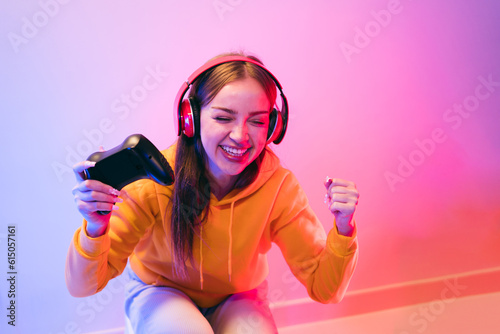 A young Caucasian beautiful woman in a yellow hoodie playing a video game using a joystick and headphones with fun, focus, surprise, and a winning victory face with blue and red neon light.