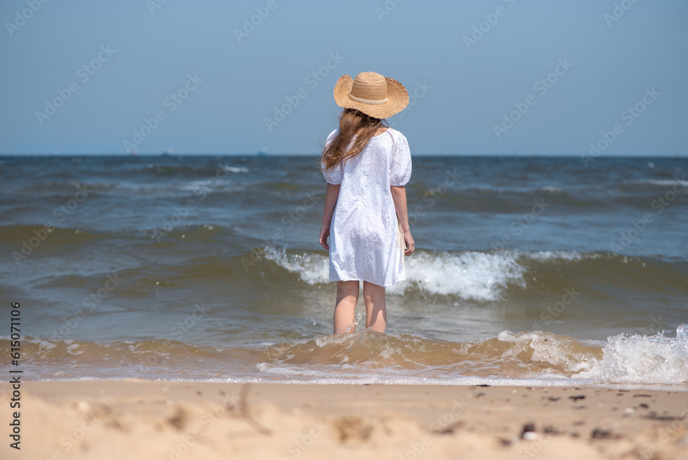 A happy girl in a white dress and a straw hat stands on the seashore, the waves wash her feet. Rear view.