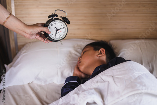 Hand of woman holding alarm clock and waking up her oversleep son at morning
