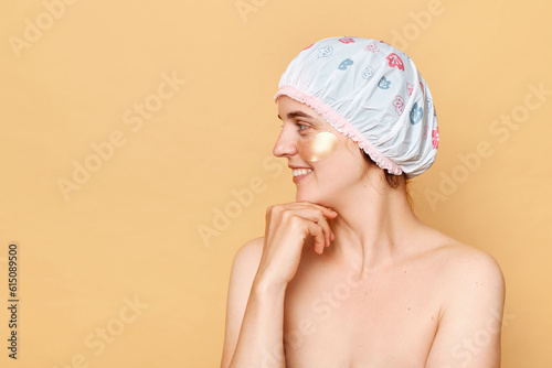 Smiling happy woman wearing shower cap with eye patches isolated over beige background looking away at copy space for advertisement, space for promotional text.