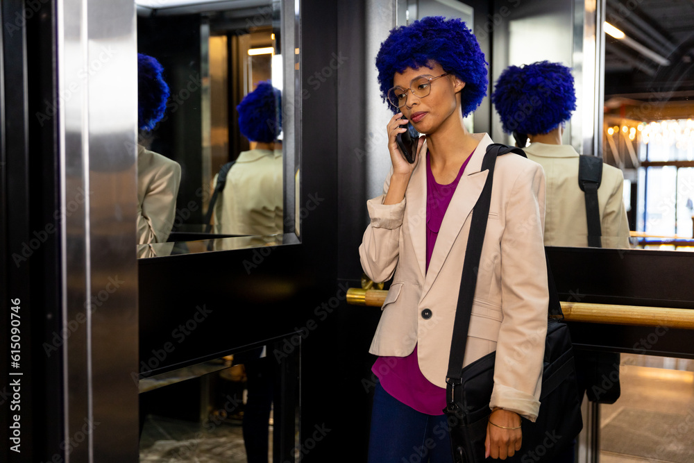 Happy biracial casual businesswoman with blue afro talking on smartphone in office elevator Happy biracial casual businesswoman with blue afro talking on smartphone in office elevator