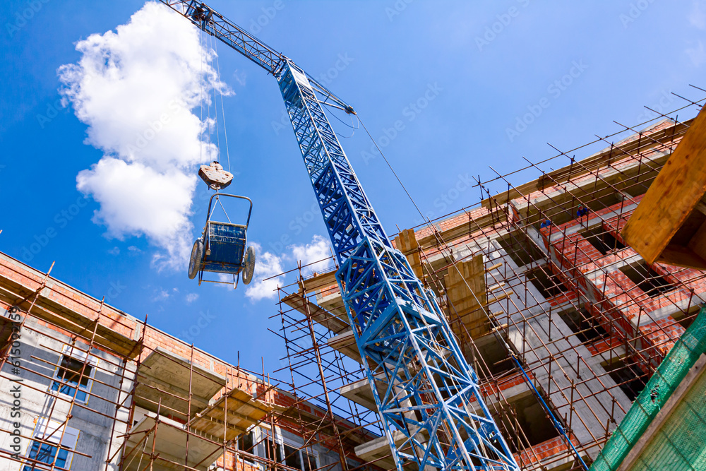 Crane lifts wheelbarrow full of mortar, in background is edifice under construction, building site