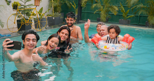 Happy Indian family fun enjoy outdoor picnic teen adult boy take selfie show thums up with parents using mobile smartphone at blue water park cute child relax in colorful floating ring tube at resort photo