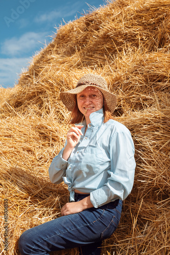 A woman in a shirt, jeans and a straw hat leans on a haystack and chews a spikelet © Neveditsyna Elena