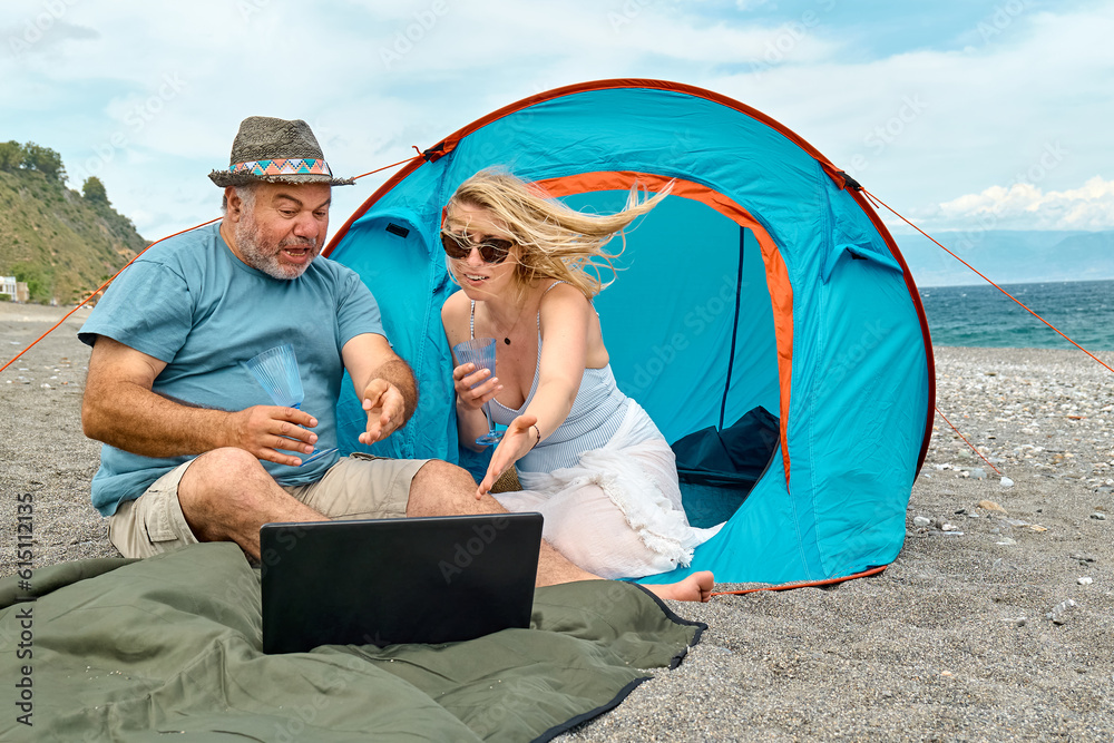 Couple having fun, drinking wine and cheer the match video on laptop during picnic near tent by the sea. Summer beach vacation outdoors.