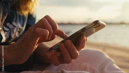 Close-up macro footage of aged female hands holding cell phone and one finger texting message social media against golden light on sunset at seaside. Elderly lady tapping screen of mobile phone. 