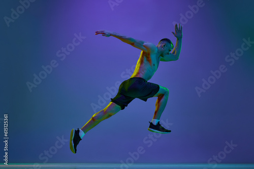 Shirtless man, with fit, relief, muscular body, professional athlete in motion, running against blue studio background in neon light