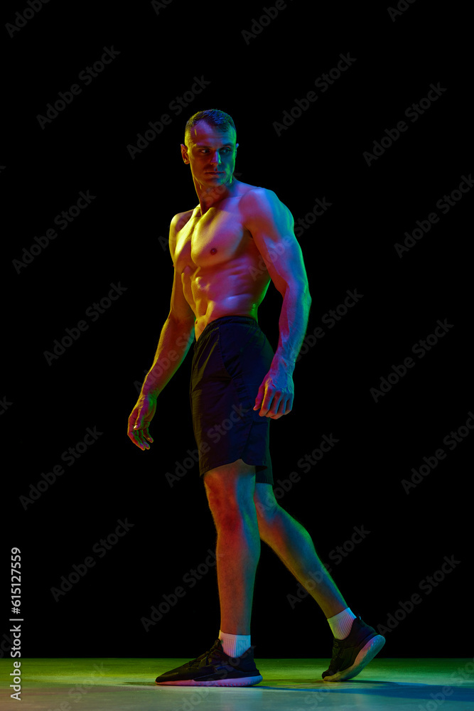 Handsome man with muscular, relief, fit, shirtless body posing against black studio background in neon light