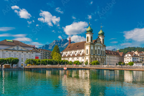 Picturesque view of the Lucerne Jesuit Church St. Francis Xavier at the river Reuss with the bridge Rathaussteg in front and the mountains in the back on a sunny day with a blue sky.