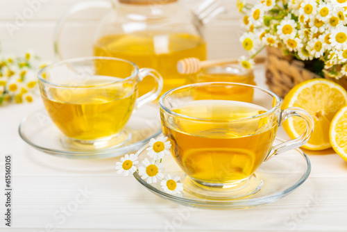 Chamomile herbal tea in a glass cup on a white wooden table with honey, lemon and chamomile bouquet. Close-up. Copy space. Useful herbal drinks, immunity tea. Natural healer concept.Place for text.
