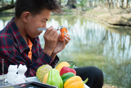 Young asian boy in plaid shirt sitting by the pond in the backyard and spending his free time by drawing, coloring and fixing his fruit and animal models sculpted from plaster with watercolor happily. #615167574