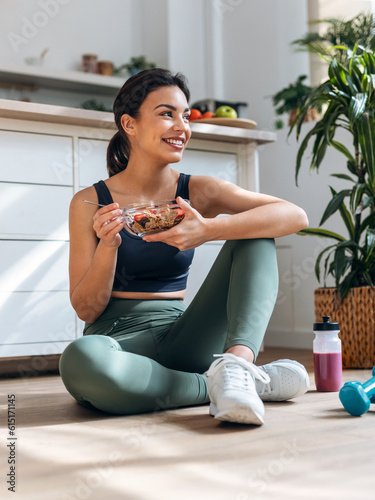 Athletic woman eating a healthy bowl of muesli with fruit sitting on floor in the kitchen at home