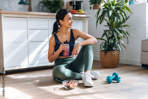 Athletic woman drinking healthy protein shake while sitting on floor in the kitchen at home