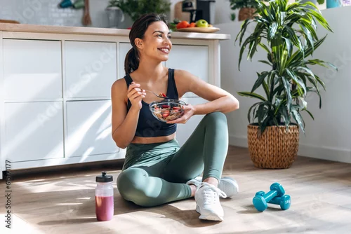 Obraz Athletic woman eating a healthy bowl of muesli with fruit sitting on floor in the kitchen at home