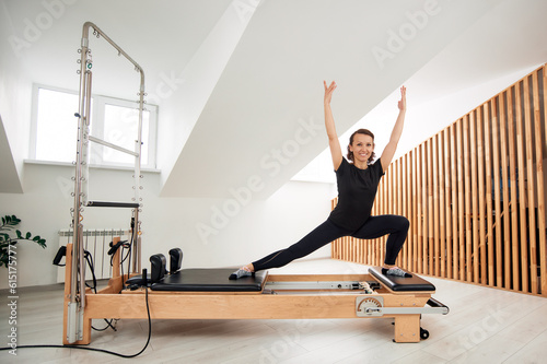 A woman is doing Pilates on a reformer bed in a bright studio. A slender flexible brunette in a black bodysuit is doing exercises to stretch the muscles. The concept of a healthy lifestyle.