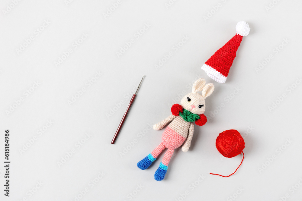 Knitted toy bunny in a red hat and scarf with ball of thread and knitting hook on gray background. Baby stuff and accessories. Gift for winter holidays. Top view, flat lay. Knitted toy bunny in a red hat and scarf with ball of thread and knitting hook on gray background. Baby stuff and accessories. Gift for winter holidays. Top view, flat lay.