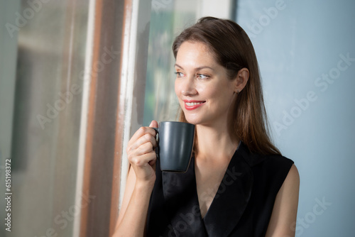Smiling beautiful business woman looking to outside office. She holding coffee cup in hand with smiling.
