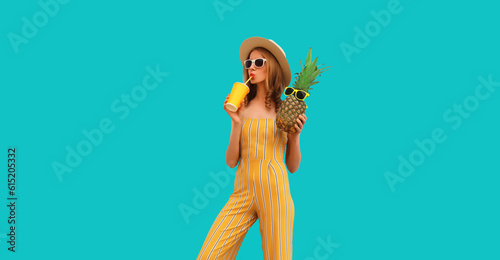 Summer portrait of young woman drinking fresh juice and holding pineapple wearing straw hat, sunglasses on blue background