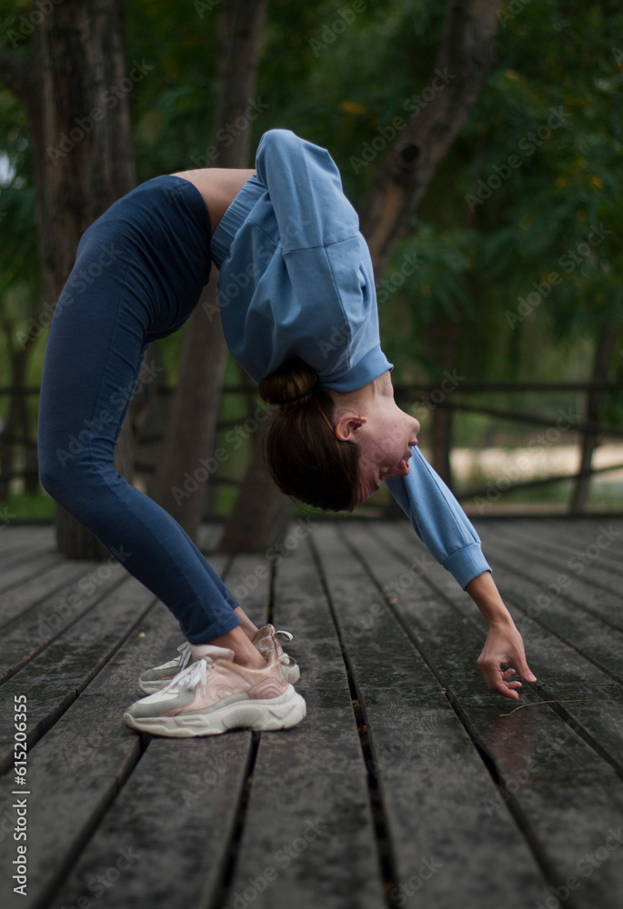 Young girl with athletic body is doing acrobatics exercises in rainy cloudy weather in the park