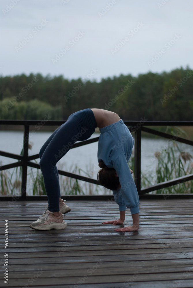 View of a girl with athletic body in blue gym suit is doing acrobatics exercises in cloudy rainy weather near lake.