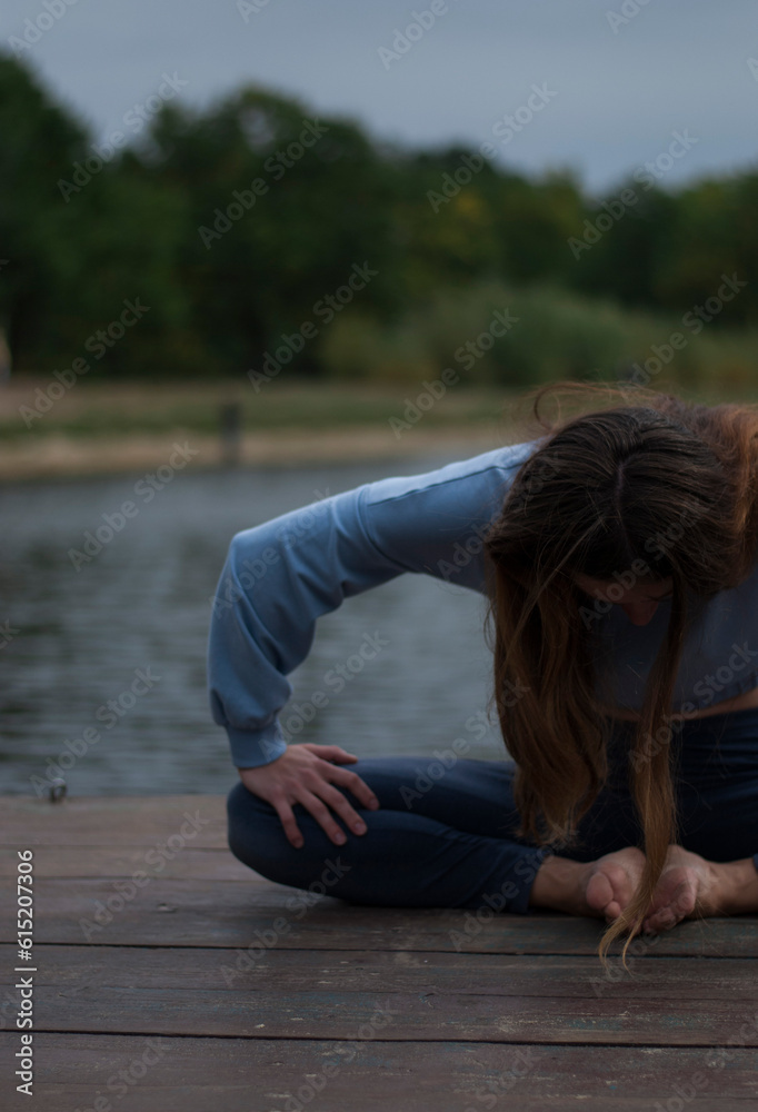 View of a girl with athletic body in  gym suit is doing yoga exercises in cloudy rainy weather near lake.