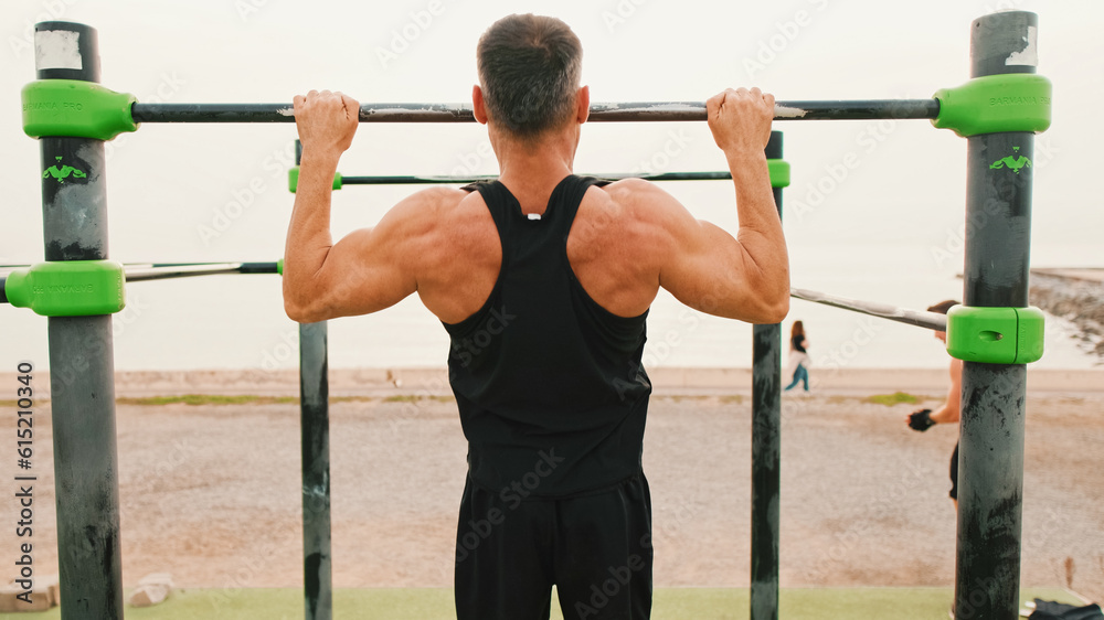 Middle-aged muscular man in sportswear pulls himself up on the horizontal bar on sea background, Back view Middle-aged muscular man in sportswear pulls himself up on the horizontal bar on sea background, Back view