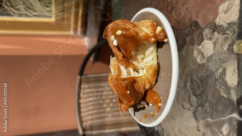 Close-up of a delicious cruffin with salted caramel torn into halves, caramel dripping down, on a saucer at a glass table of a cafe terrace. Lifestyle vertical video. photo