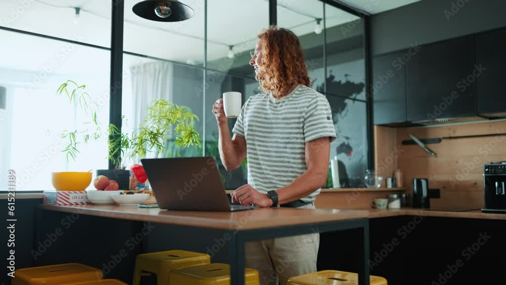 Happy curly haired man in eyeglasses drinking cup of coffee after working on laptop in office kitchen Happy curly haired man in eyeglasses drinking cup of coffee after working on laptop in office kitchen