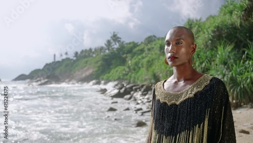 Queer black person in luxury dress, handmade jewelry walks on scenic sea cliff. Closeup slow motion. Lgbtq biethnic fashion gay model in posh gown, brass accessories portrait. Tropical location. photo