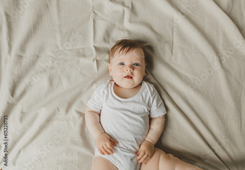Charming blue-eyed 6-month-old baby lies in bed in a white bodysuit. View from above
