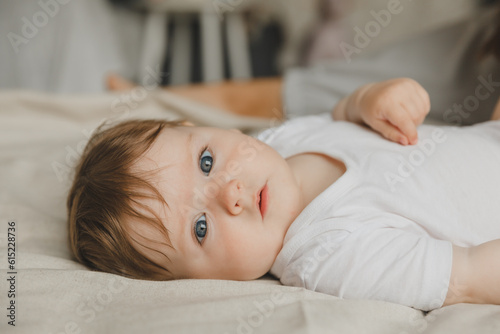 Fotografija Charming blue-eyed 6-month-old baby lies in bed in a white bodysuit