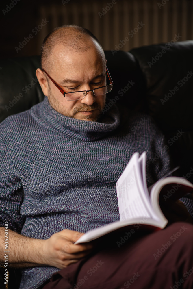 A man with glasses is reading a book. Middle-aged man of ordinary appearance in casual clothes at home. Reading, learning.