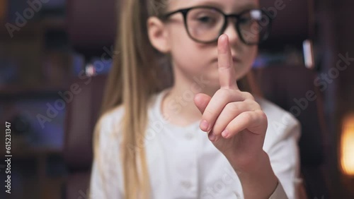 Crop of serious small girl looking at camera and waving index finger to say no. Clever preteen kid in glasses sitting in rolling office chair, shaking head and making denial gesture.