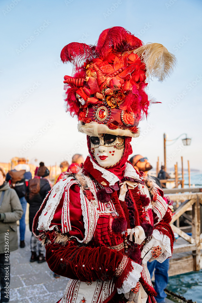 Venice, Italy, 11 February 2023: Colorful carnival masks at famous traditional festival on Saint Mark's Square at sunset, Elegant Venetian Costume, red velvet dress with embroidery and feathers