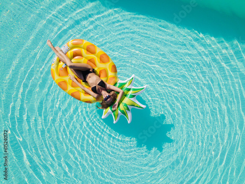 Aerial view of female in bikini lying on a floating mattress in swimming pool with her face covered with hat. Top view of woman sunbathing on inflatable mattress in pool.