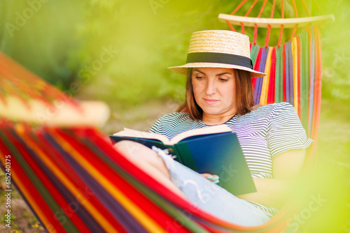 Young woman reading book while lying in comfortable hammock at green garden