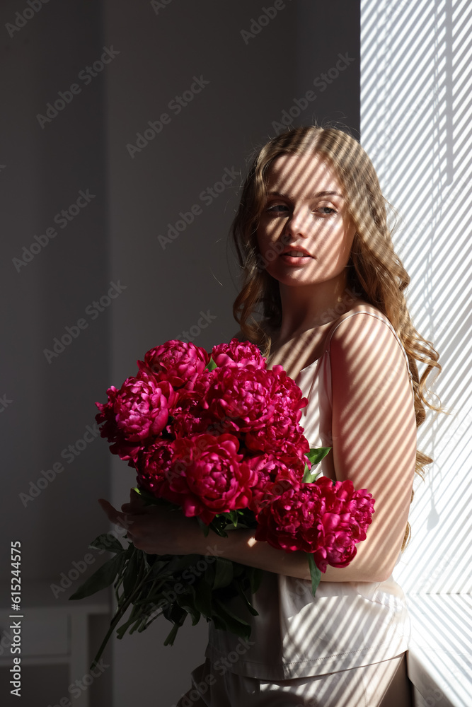 Beautiful young woman with bouquet of peony flowers near window with blinds Beautiful young woman with bouquet of peony flowers near window with blinds