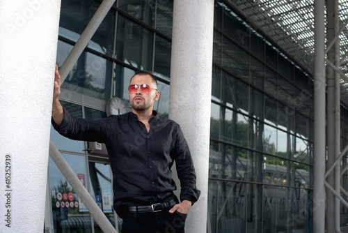 a stylish man in light pants and a black shirt against the background of empty walls holds glasses in his hands and looks away angrily