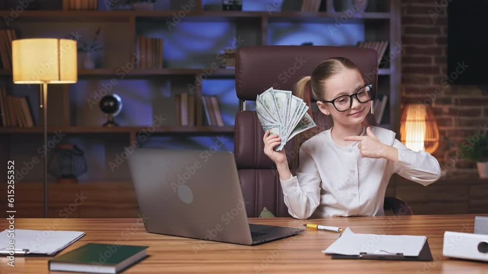 Petite business girl posing on camera in front of laptop and pointing at bunch of dollar banknotes. Pretty kid enthusiastically nodding head with raised eyebrows. Concept of huge financial profit.