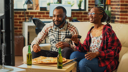 Cheerful man and woman eating slices of pizza at home, being relaxed together watching movie on television. Young people in relationship enjoying delivery meal and beer bottles.