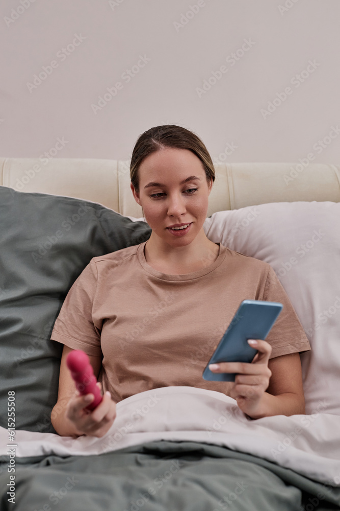 Vertical portrait of young woman holding sex toy and smartphone while sitting in bed at home Vertical portrait of young woman holding sex toy and smartphone while sitting in bed at home