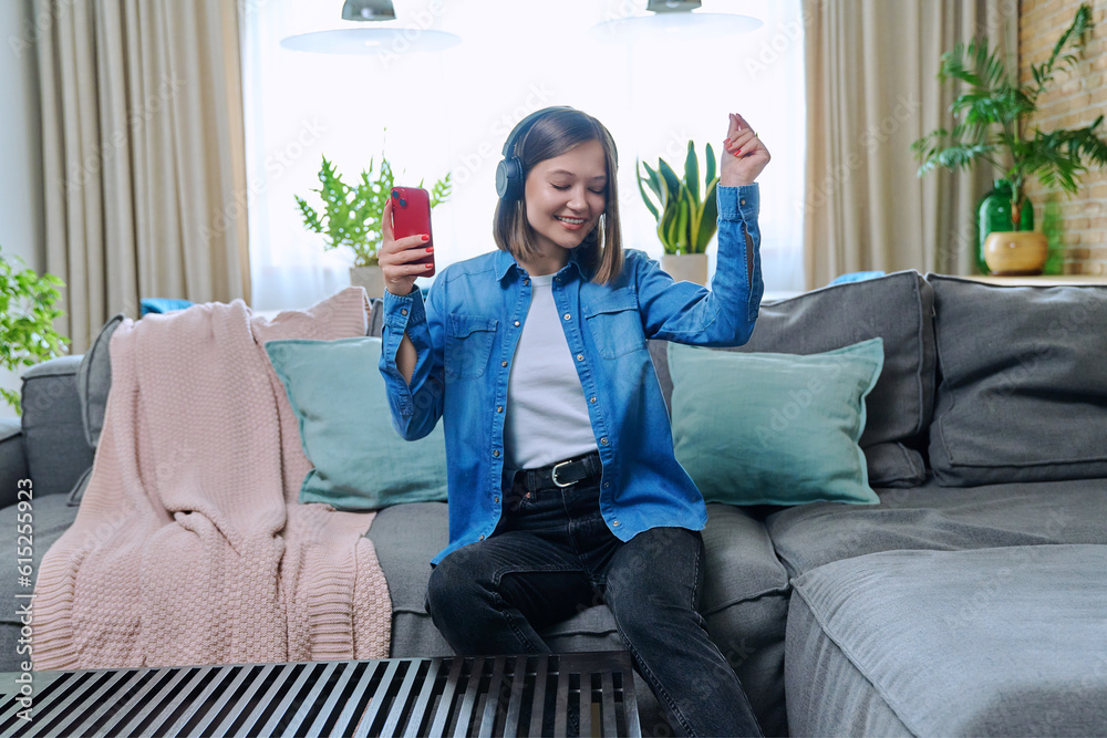 Obraz premium Young woman in headphones enjoying music, dancing, sitting on sofa at home
