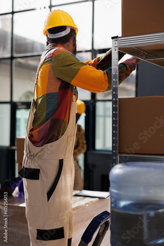 African american man standing on ladder and searching parcel on shelf in industrial warehouse. Storehouse worker wearing hard hat taking cardboard box for client order packing in stockroom