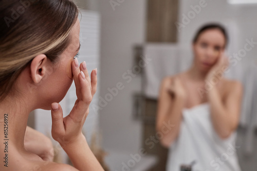 Closeup of young woman using eye cream in bathroom and looking in mirror enjoying self care day at home, copy space