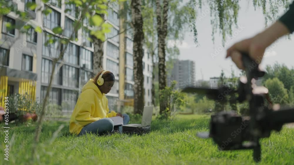 Shooting behind scenes. Shooting video with professional camera. Dark-skinned model girl is resting on lawn in courtyard of residential complex. Backstage. Slow motion Shooting behind scenes. Shooting video with professional camera. Dark-skinned model girl is resting on lawn in courtyard of residential complex. Backstage. Slow motion